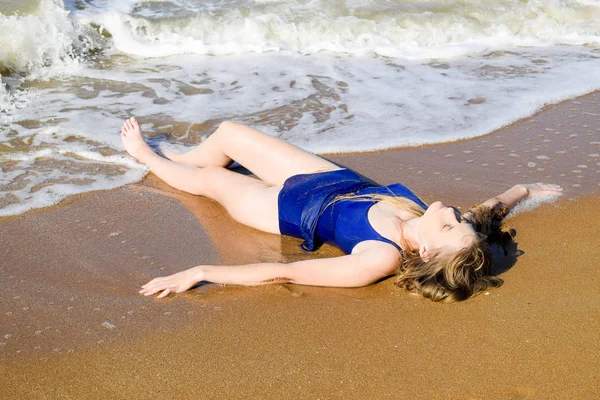 Girl in a blue bathing suit lies on the seashore and relaxes. Waves caress the girls body — Stock Photo, Image