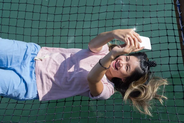 Mujer asiática sosteniendo el teléfono, sonriendo y acostada en la hamaca — Foto de Stock