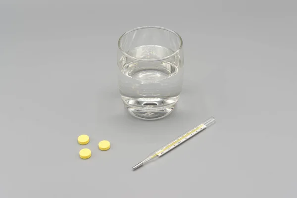 Pílulas amarelas, termômetro e vidro de água em fundo cinza — Fotografia de Stock