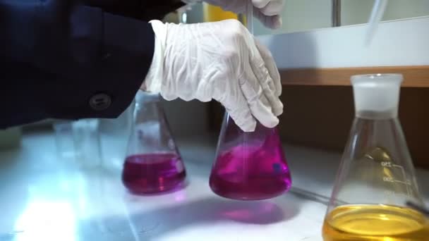 Hands in gloves mixes pink liquid and yellow liquid in flask in lab — Stock Video