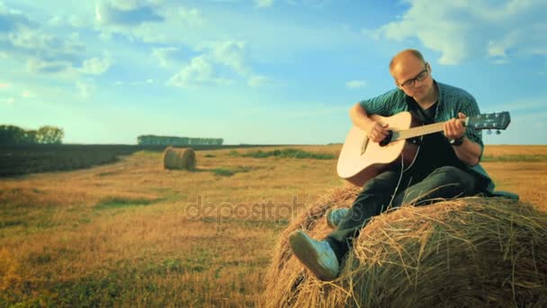 Mujer bastante joven tocando la guitarra, sentado contra un rollo de heno . — Vídeos de Stock