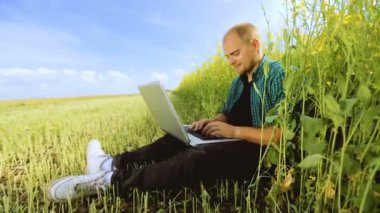 Çimenlerin üzerinde oturan ve dizüstü bilgisayarda yazarak gözlüklü genç adam