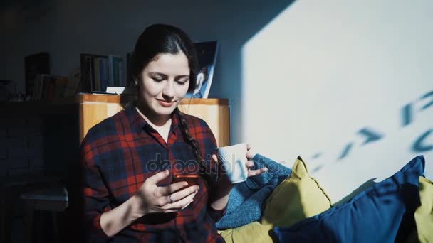 Een jonge vrouw zit in een café en teksten op haar mobiele telefoon terwijl het drinken van een kopje koffie — Stockvideo