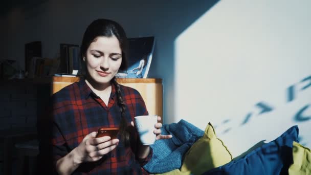 Una giovane donna si siede in un caffè e scrive sul suo cellulare mentre beve una tazza di caffè — Video Stock