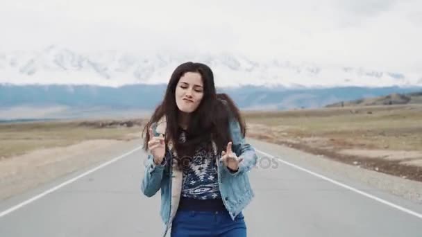 Linda menina adolescente hipster dançando na estrada contra o pano de fundo das montanhas. Estou feliz e feliz. Jaqueta de jeans e morena — Vídeo de Stock