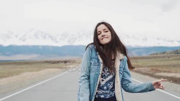 Mooie tiener meisje hipster dansen op de weg tegen de achtergrond van de bergen. Im blij en gelukkig. Jeans jasje en brunette — Stockvideo