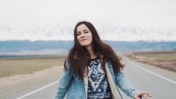 Hermosa chica adolescente hipster bailando en el camino contra el telón de fondo de las montañas. Estoy feliz y feliz. Chaqueta vaqueros y morena — Vídeo de stock
