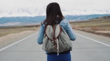 Genç kız genç hippi karayolu üzerinde yürüme. Kamp bir sırt çantası ve bir jersey ceket ile. Karlı dağlar fonunda. Kız yalnız seyahat