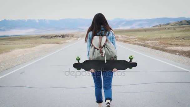 Fiatal lány tizenéves csípő sétál a longboard autópálya. A kemping hátizsák és egy jersey kiskabát. Havas hegyek a háttérben. A lány egyedül utazik — Stock videók