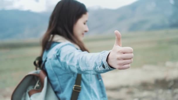 Mujer joven haciendo autostop en una carretera en los campos. Niña hippy haciendo autostop en la carretera. Entre las montañas en la naturaleza. Voy a la carretera. Primer plano de un pulgar — Vídeos de Stock