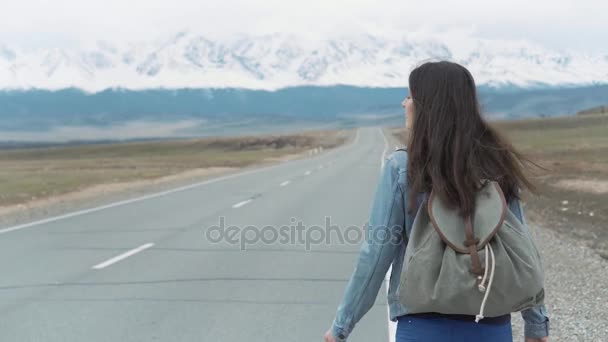 Eine junge Frau trampt auf einem Feldweg. Hippie-Mädchen trampen auf der Straße. zwischen den Bergen in der Natur. Unterwegs. — Stockvideo