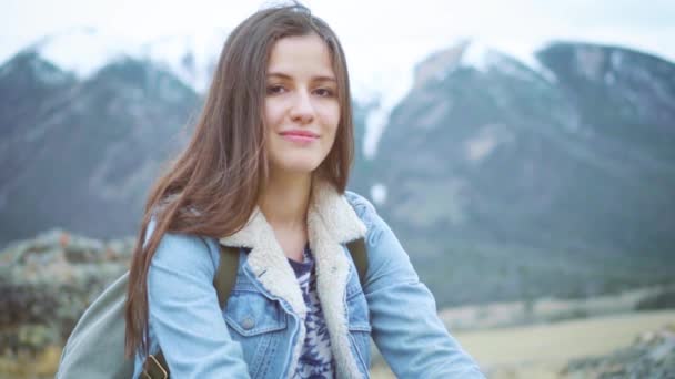 Ung flicka resenären sitter på klippan, njuter av utsikten över bergen, ler mot kameran — Stockvideo