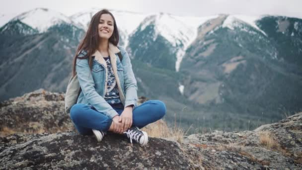 Молодая девушка путешественник сидит на скале, наслаждаясь видом на горы, улыбаясь в камеру — стоковое видео