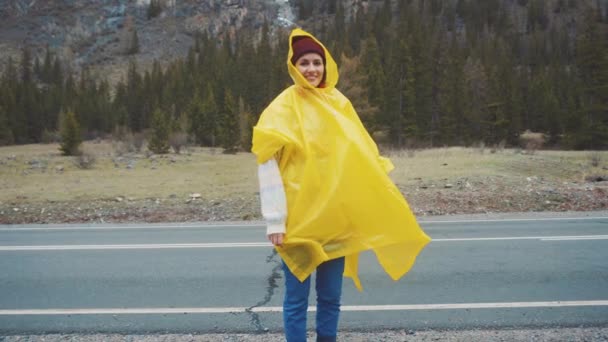 Πορτρέτο: ένα όμορφο κορίτσι στέκεται σε ασφαλτοστρωμένο δρόμο ή εθνική οδό τον χειμώνα με φόντο τα βουνά. Απολαμβάνει ταξιδεύουν ή περιπέτεια. Ντυμένος με ένα κίτρινο αδιάβροχο — Αρχείο Βίντεο
