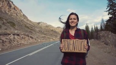 Genç kız bir macera işareti Holding bir dağ yolu, gülümsüyor ve gülüyor. Macera arayışı içinde otostop.