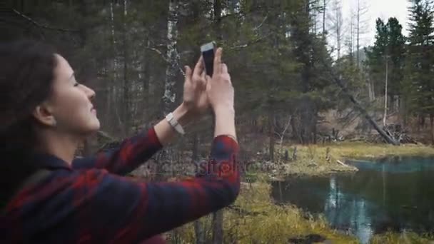 Happy girl traveler senta-se na rocha, faz uma foto no lago cratera de montanha smartphone. Vestida com um casaco de jeans e mochila para caminhadas. Viaje sozinho, encontrando-se . — Vídeo de Stock