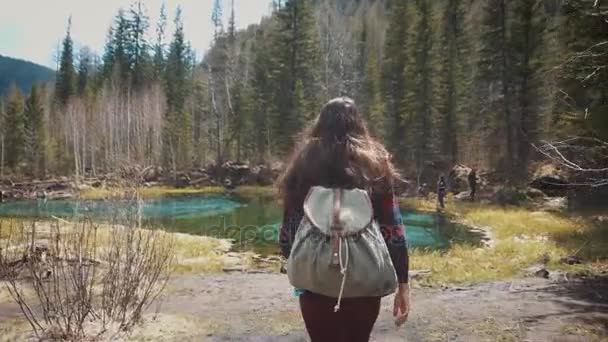 Woman hiker walking around mountain lake and enjoys his spectacular sceneries. On a background of a dense pine forest. Dressed in a jeans jacket and hiking backpack. Travel alone, finding yourself. — Stock Video