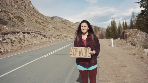 Дівчина-підліток тримає знак пригоди на гірській дорозі, посміхається і сміється. Автостоп у пошуках пригод . — стокове відео