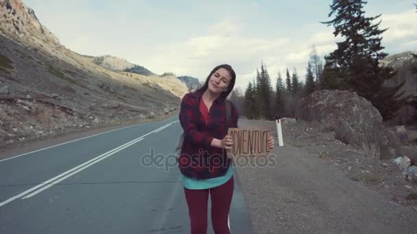 Adolescente segurando um sinal de aventura em uma estrada da montanha, sorrisos e gargalhadas. Carona em busca de aventura . — Vídeo de Stock