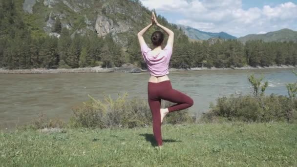 Young beautiful woman in red tank top practicing outdoors, doing Bending Tree posture, variation of Vrksasana, in park on summer day. Altai, the river katun. — Stock Video