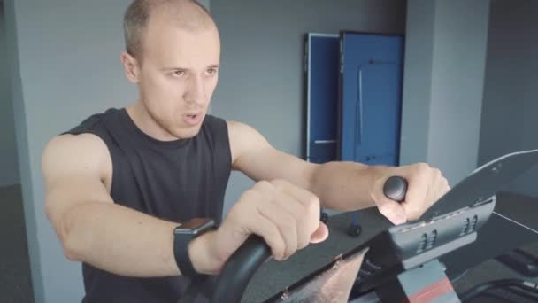 Молодий чоловік зі смарт-годинником їде на стаціонарному велосипеді у спортзалі — стокове відео