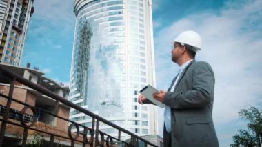 Yetişkin mühendis veya mimar bir tablet işlemi kullanır. Bir ileti yazar veya bir çizim denetler. Arka planı yapıdır. İnşaatçılar beton ve cam modern konut bina inşa ediyoruz