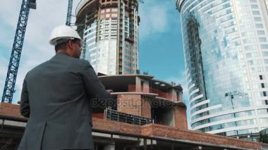 Yetişkin mühendis veya mimar bir tablet işlemi kullanır. Bir ileti yazar veya bir çizim denetler. Arka planı yapıdır. İnşaatçılar beton ve cam modern konut bina inşa ediyoruz