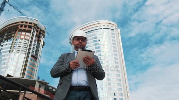 Adult engineer or architect uses a tablet in operation. Writes a message or checks a drawing. Against background is building. Builders are building a modern residential building of glass and concrete — Stock Video