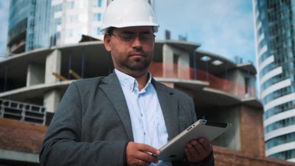 Yetişkin mühendis veya mimar bir tablet işlemi kullanır. Bir ileti yazar veya bir çizim denetler. Arka planı yapıdır. İnşaatçılar beton ve cam modern konut bina inşa ediyoruz — Stok video