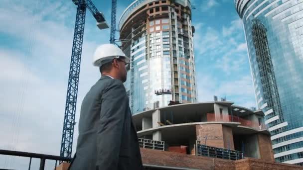 O engenheiro ou construtor verifica e controla o objeto em construção. Ele passa pelo estaleiro, os trabalhadores estão a trabalhar. Contra o pano de fundo de edifícios modernos inovadores — Vídeo de Stock