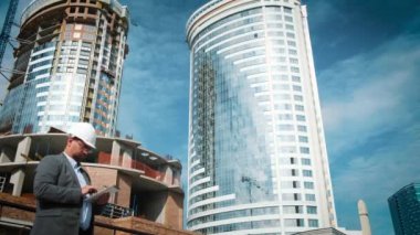 Yetişkin mühendis veya mimar bir tablet işlemi kullanır. Bir ileti yazar veya bir çizim denetler. Arka planı yapıdır. İnşaatçılar beton ve cam modern konut bina inşa ediyoruz