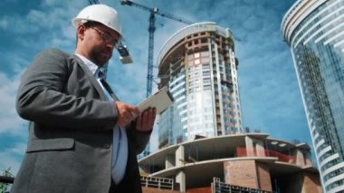 Yetişkin mühendis veya mimar bir tablet işlemi kullanır. Bir ileti yazar veya bir çizim denetler. Arka planı yapıdır. İnşaatçılar beton ve cam modern konut bina inşa ediyoruz