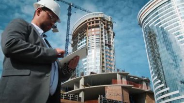 Yetişkin mühendis veya mimar bir tablet işlemi kullanır. Bir ileti yazar veya bir çizim denetler. Arka planı yapıdır. İnşaatçılar beton ve cam modern konut bina inşa ediyoruz
