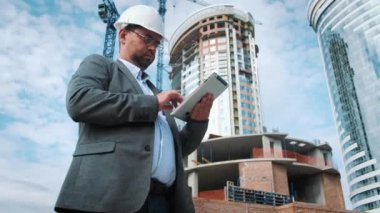 Yetişkin mühendis veya mimar bir tablet işlemi kullanır. Bir ileti yazar veya bir çizim denetler. Arka planı yapıdır. İnşaatçılar beton ve cam modern konut bina inşa ediyoruz