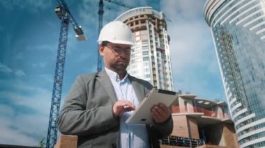 Yetişkin mühendis veya mimar bir tablet işlemi kullanır. Bir ileti yazar veya bir çizim denetler. Arka planı yapıdır. İnşaatçılar beton ve cam modern konut bina inşa ediyoruz