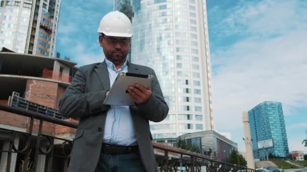 Ingeniero adulto o arquitecto utiliza una tableta en funcionamiento. Escribe un mensaje o comprueba un dibujo. Contra el fondo se está construyendo. Los constructores están construyendo un moderno edificio residencial de vidrio y hormigón — Vídeo de stock