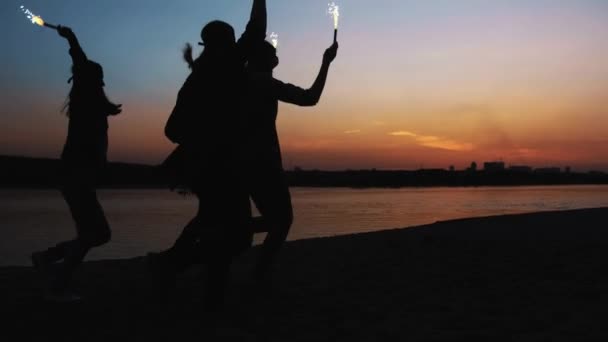 Retrovisore di amici che corrono insieme su una spiaggia con fuochi d'artificio scintillanti. fidanzate fidanzate corrono lungo la riva di sera o di notte — Video Stock