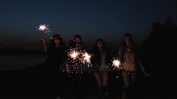 Visão traseira de amigos correndo juntos em uma praia segurando fogos de artifício cintilantes. namoradas namoradas correr ao longo da costa à noite ou à noite — Vídeo de Stock