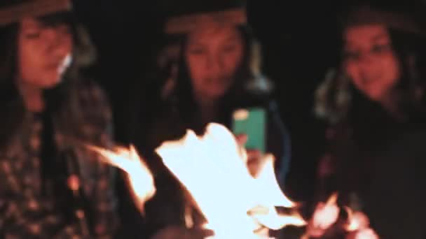 Plusieurs filles heureuses sont assises à côté d'un feu de camp la nuit et utilisent des smartphones. Ils regardent la photo, sourient. Correspondre avec les gars . — Video