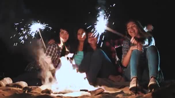 Picnic of young people with bonfire on the beach in the evening. Cheerful friends singing songs and playing guitar. Hold the burning Bengal lights in their hands. — Stock Video