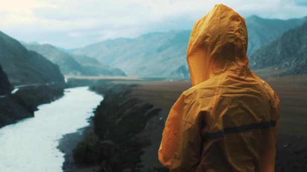 在雨季，年轻女孩看着山景观。女人穿着黄色的雨衣。徒步旅行者是背包。她享受美丽的自然景观. — 图库视频影像