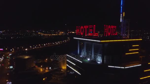 AÉRIAL : Hôtel inscription rouge fluo sur le toit d'un bâtiment moderne dans la ville nocturne. Les voitures de circulation sont les prochaines sur la route. Le mot hôtel sur le toit d'un gratte-ciel — Video