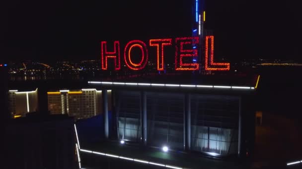 空中: 霓虹灯红色题字旅馆在一个现代大厦的屋顶在夜城市。道路上的交通车辆是下一个。摩天大楼顶上的那个词酒店 — 图库视频影像