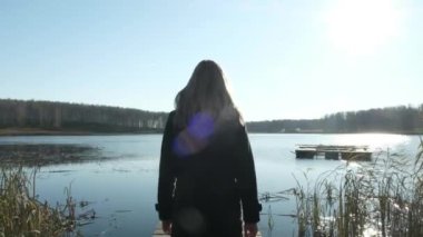 Güzel sarışın kız paltolu bir iskelede göl kenarında yürüyor. Göl, sonbahar ve bulutlu ruh hali