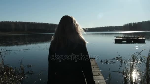 Een mooie blonde meisje met een vacht loopt op een pier van het meer. Op het meer, de herfst en de bewolkt stemming — Stockvideo