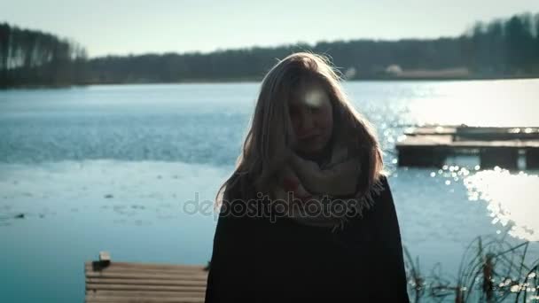 穿着大衣的漂亮金发女郎站在湖边的一个码头上。湖上, 秋天和多云的心情. — 图库视频影像