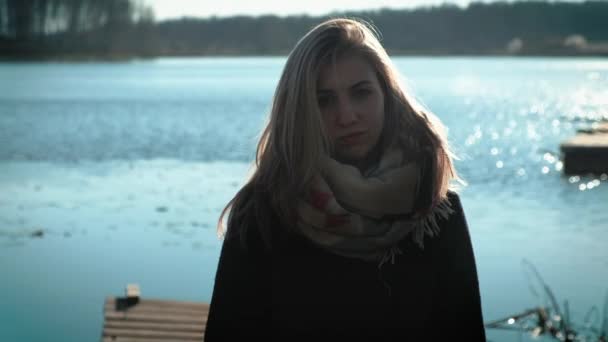 Beautiful blonde girl in a coat is standing on a pier by the lake. On the lake, autumn and cloudy mood. — Stock Video