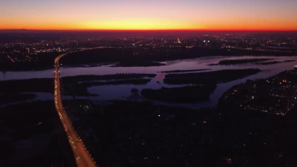 Vídeo aéreo de Novosibirsk Rusia. Ciudad nocturna al atardecer. Puesta de sol reflejada en el río. Conduce coches en el puente . — Vídeos de Stock