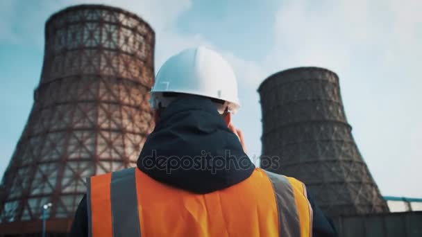 Профессиональный инженер в белом шлеме и куртке смотрит на заводские трубы. От градирен идет белый пар или дым — стоковое видео