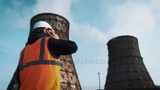 Professional engineer in a white helmet and jacket looks at the factory pipes. She calls on the phone. From the cooling towers there is white steam or smoke. — Stock Video